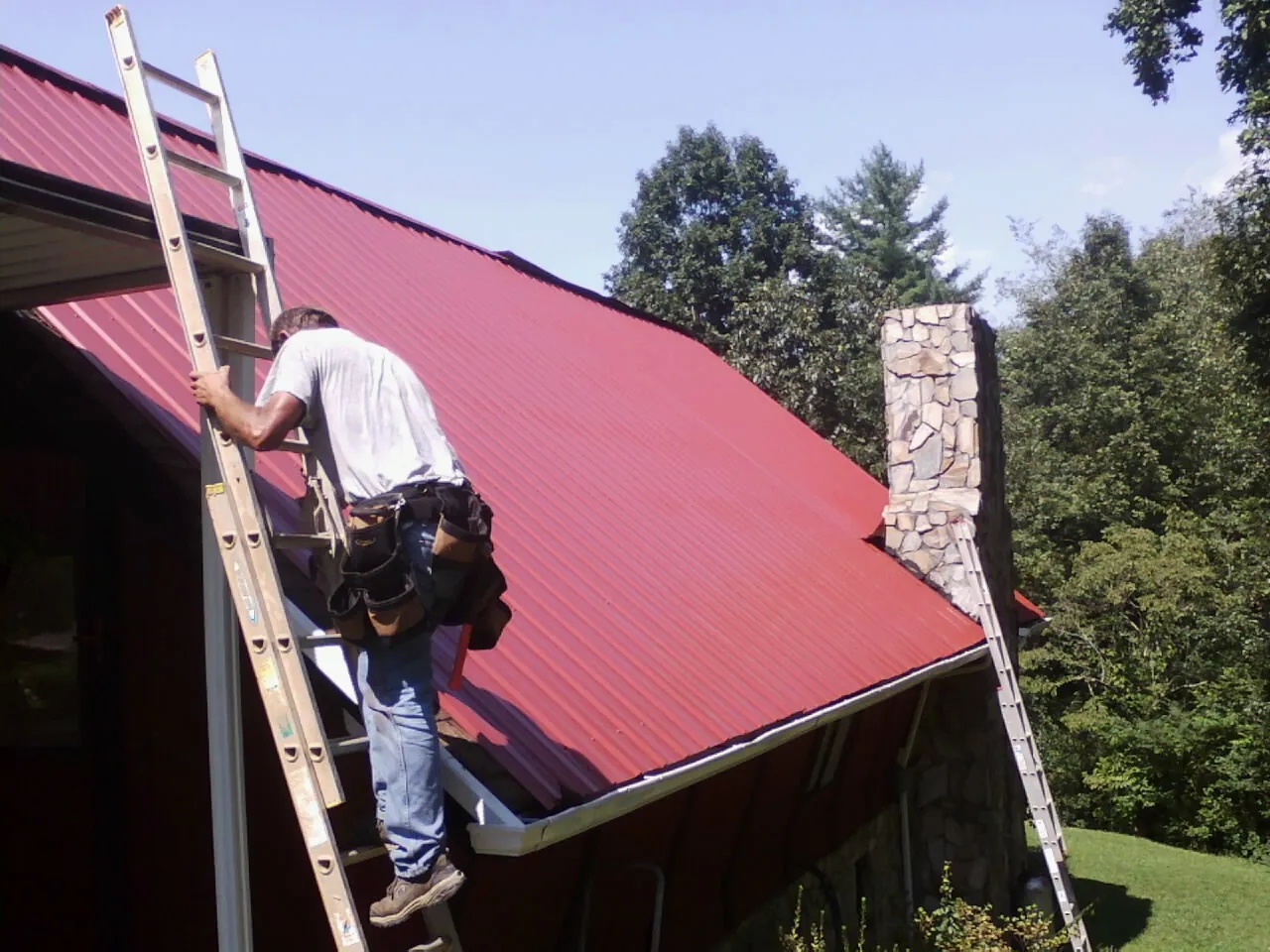 Roofer on a ladder beside a red metal roof and stone chimney — Soffit & Fascia Repair by Roofing Brothers in Kingston Springs